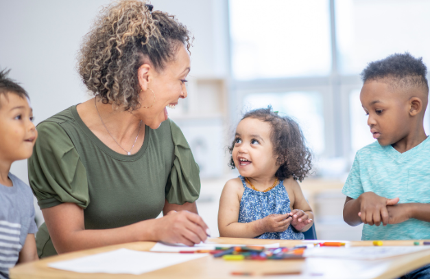 A pre-school teacher sits at a table with three young students. They are coloring with crayons, and smiling at each other.