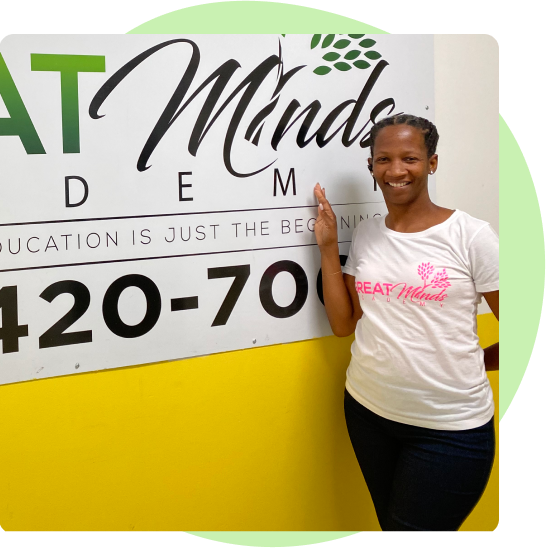 JOY BOULER stands proudly in front of a sign with the name of her early learning center on it, Great Minds Academy. She is smiling and wearing a t-shirt that also says the name of her business.