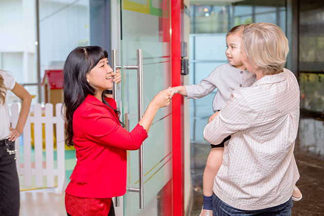 A preschool teacher greets  a parent and child at the front door of an early learning center.  The parent holds the child in their arms, and the child reaches out to shake the teacher's hand. 