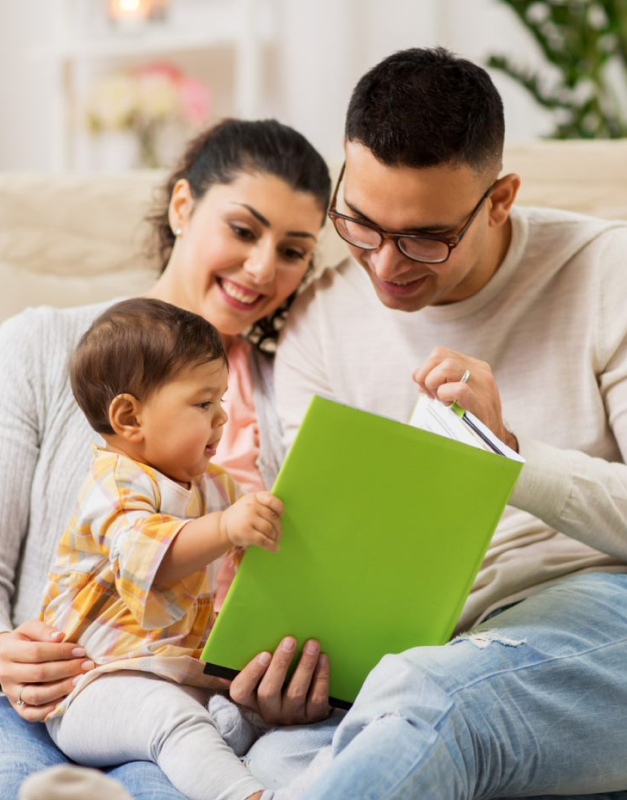 A couple sits with their infant child in their laps. One of the parents hold a book, and the infant looks at it with curiosity.