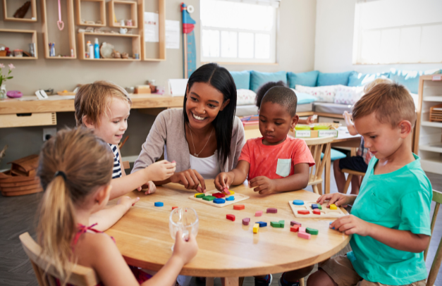a teacher sits at a table in an early learning center, surrounded by four students. The children are doing an activity, and the teacher smiles at their work.