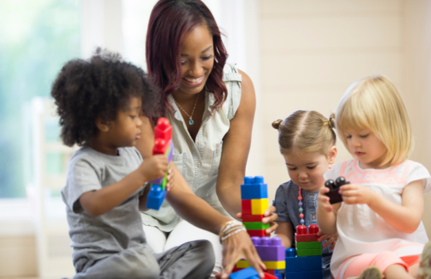 A woman sits on the floor with three young children under the age of four. The children are playing with building blocks, and the woman reaches out to offer a block to one of the children.