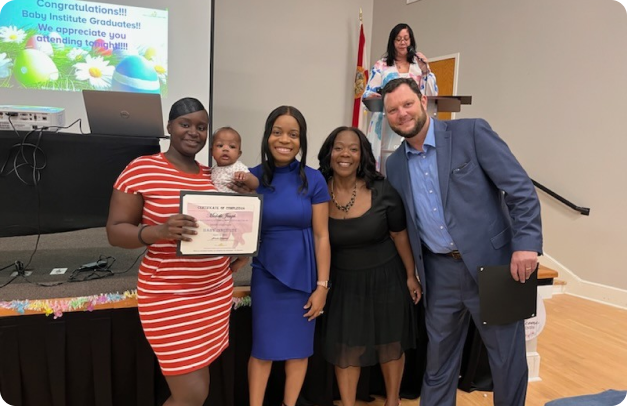 A family stands in front of a stage with their infant. They have just graduated from the Baby Institute program. All three women smile, while one holds the infant and their certificate of completion.