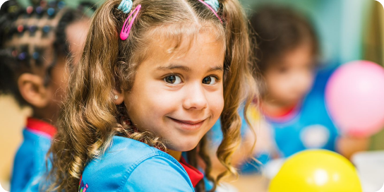 a four year old girl smiles in her pre-school classroom. She and her classmates all wear matching shirts, their school uniform.