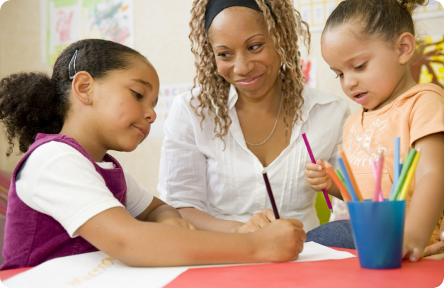 A teacher sits at a low table with two pre-school students. The students are drawing together and the teacher looks on with pride.