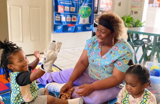 A pre-school teacher sits on the floor with two students. She is laughing, while one of the students holds a puppet. They are having an interactive conversation to develop the children's language skills.