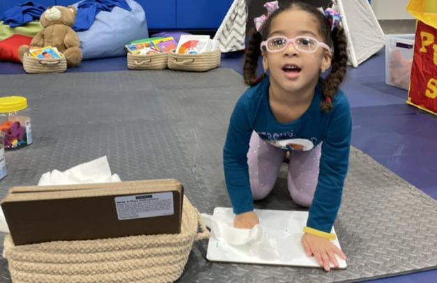 A preschool student has been doing an activity on a small, personal dry-erase board. She wipes her dry-erase board clean with a cloth.