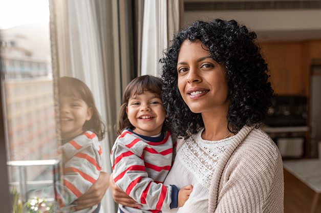 A woman stands confidently with her child on her hip. The woman and child are smiling, and the woman has dark, curly hair. The child is around 3 years old.